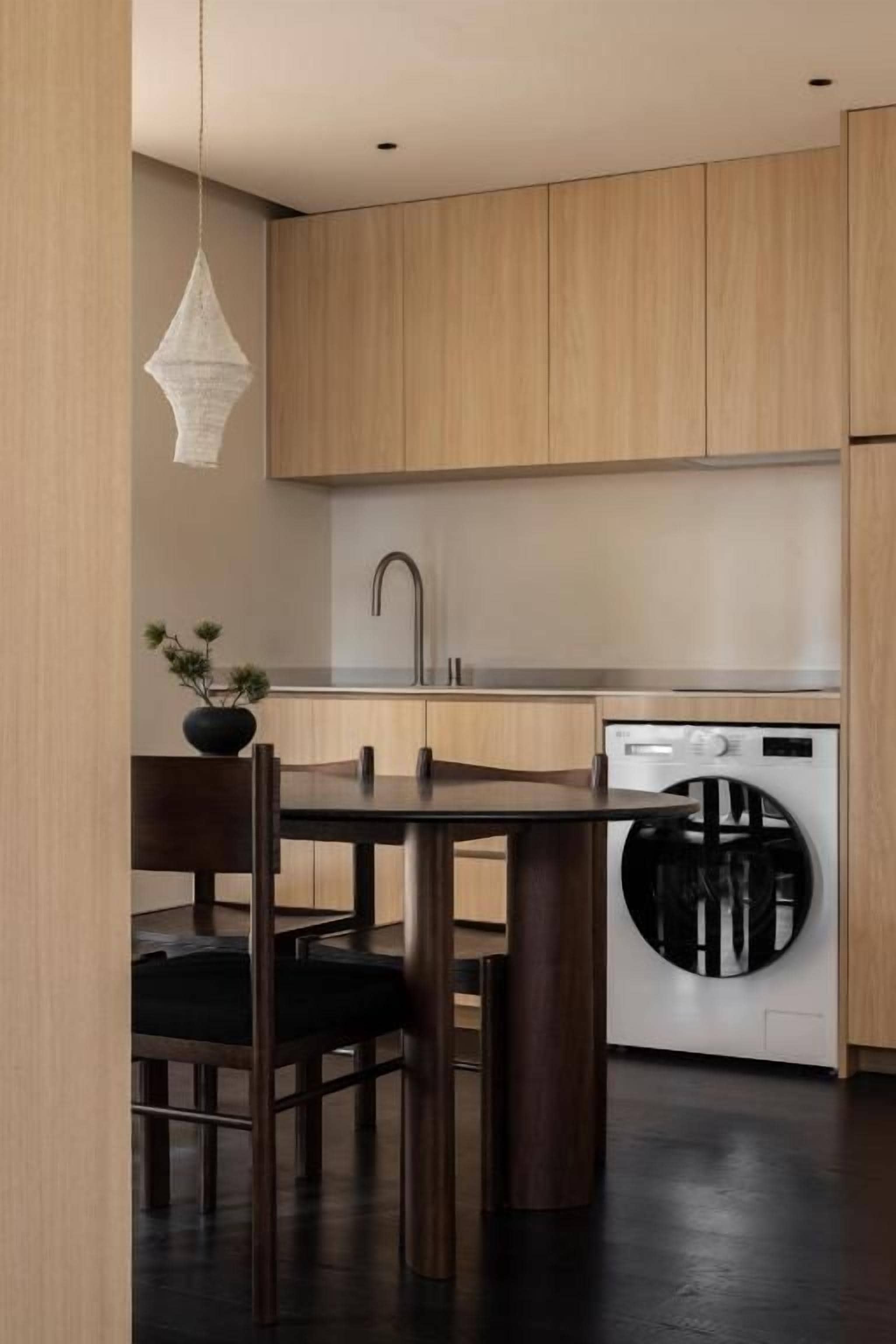 A modern kitchen with wooden cabinets, a sink, a washing machine, and a dining table with chairs.