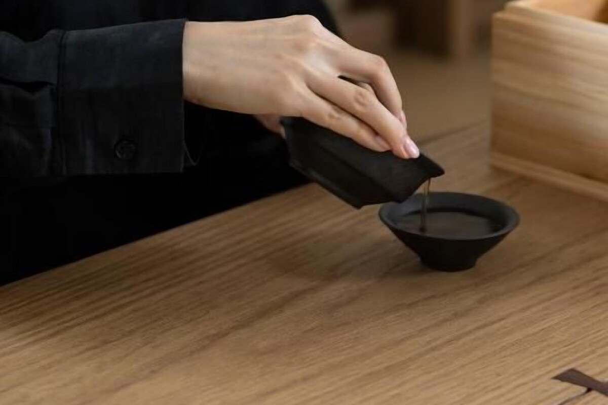 Una persona vertiendo un líquido desde un recipiente negro hacia un tazón negro sobre una mesa de madera.