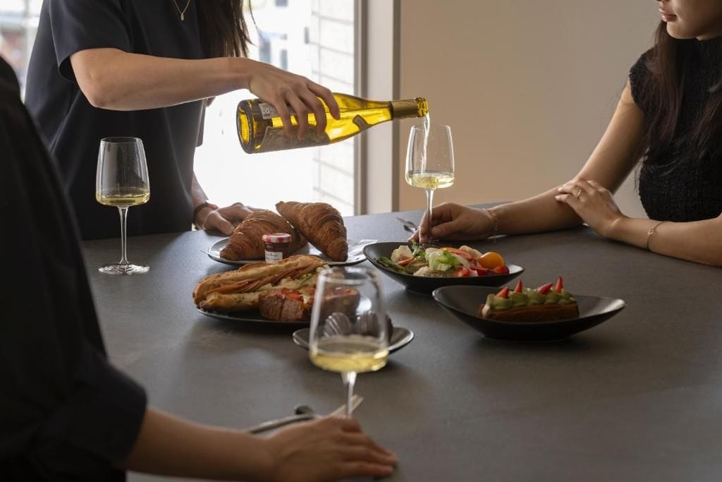 A dining table with food and wine glasses, a bottle being poured, and a person pouring wine.