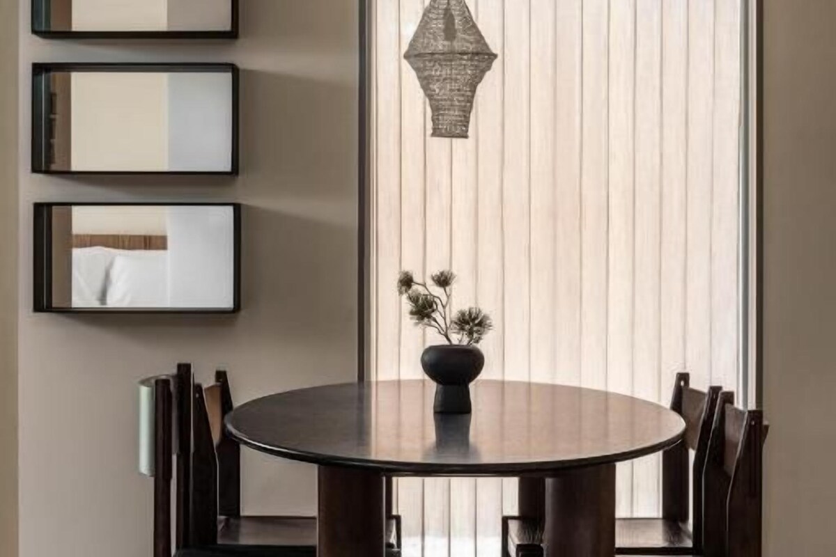 Un comedor con una mesa redonda, sillas, una lámpara colgante y una ventana con persianas.