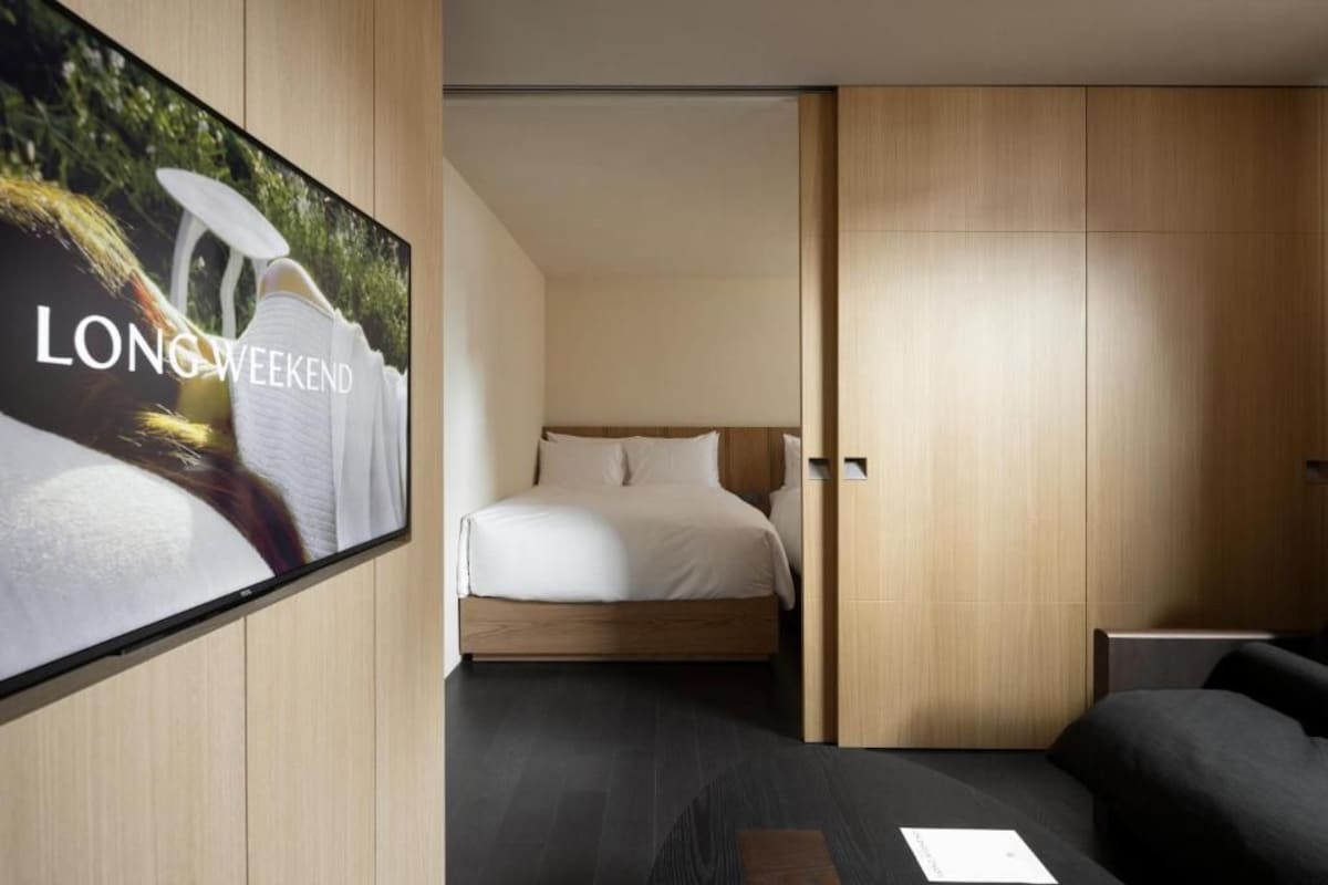 Quarto de hotel moderno com uma cama, televisão fixada na parede e uma porta de madeira deslizante.