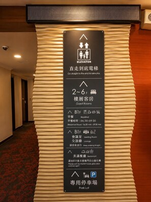 Interior de un hotel con un cartel direccional que indica la ubicación del ascensor, habitaciones de huéspedes, desayuno, sala de reuniones, salón, restaurante y estacionamiento.