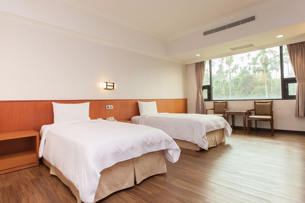 Habitación de hotel con dos camas, piso de madera, ventana con vistas a palmeras y un escritorio con silla.