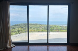 Una habitación con un gran ventanal corredera que da a un balcón con vista al océano y al follaje.