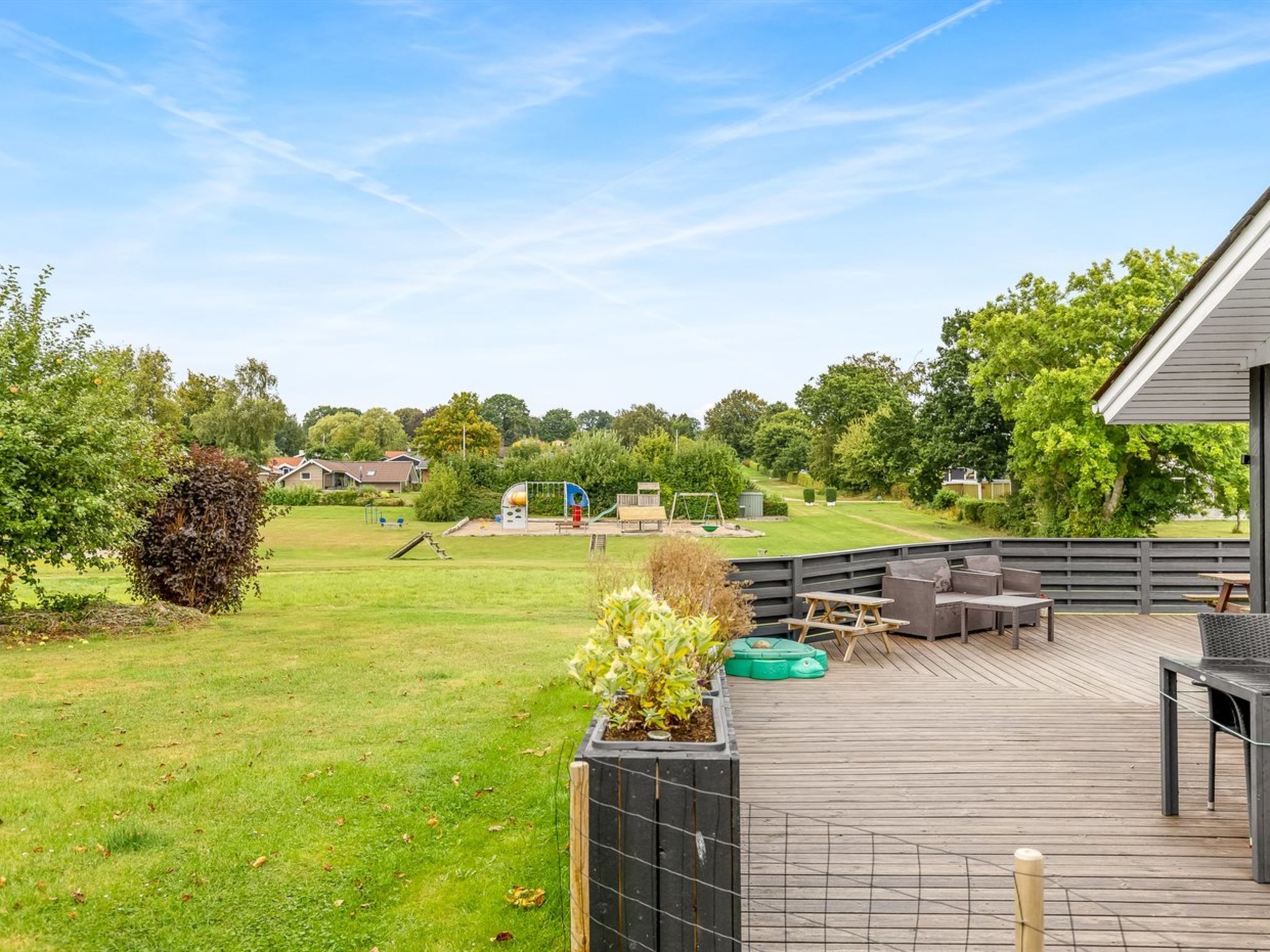 A wooden deck with outdoor furniture, a grassy yard, and a playground in the background.
