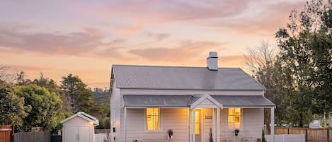 Une maison de plain-pied avec un toit à pignon, un porche à l’avant avec deux chaises et un petit abri de jardin sur le côté gauche.