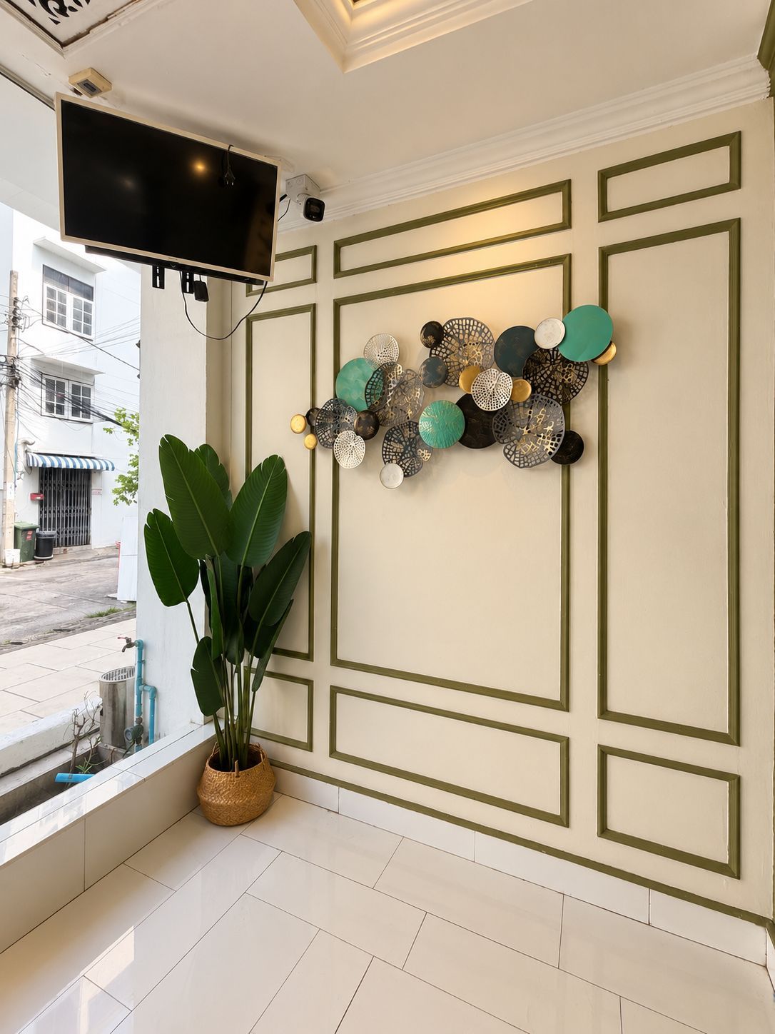 A room with a flat-screen TV mounted on the wall, a decorative wall art piece, a large potted plant, and a window with a view of the street.