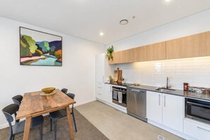 A modern kitchen with white cabinetry, a wooden dining table, and a colorful abstract painting on the wall. - Christchurch CBD - 2BR with Parking & Balcony (Christchurch)