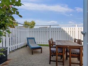 An outdoor patio with a table and chairs, a lounge chair, and a white fence.