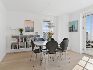 Uma área de refeições com uma mesa branca, cadeiras pretas, uma estante de livros e um janelão.