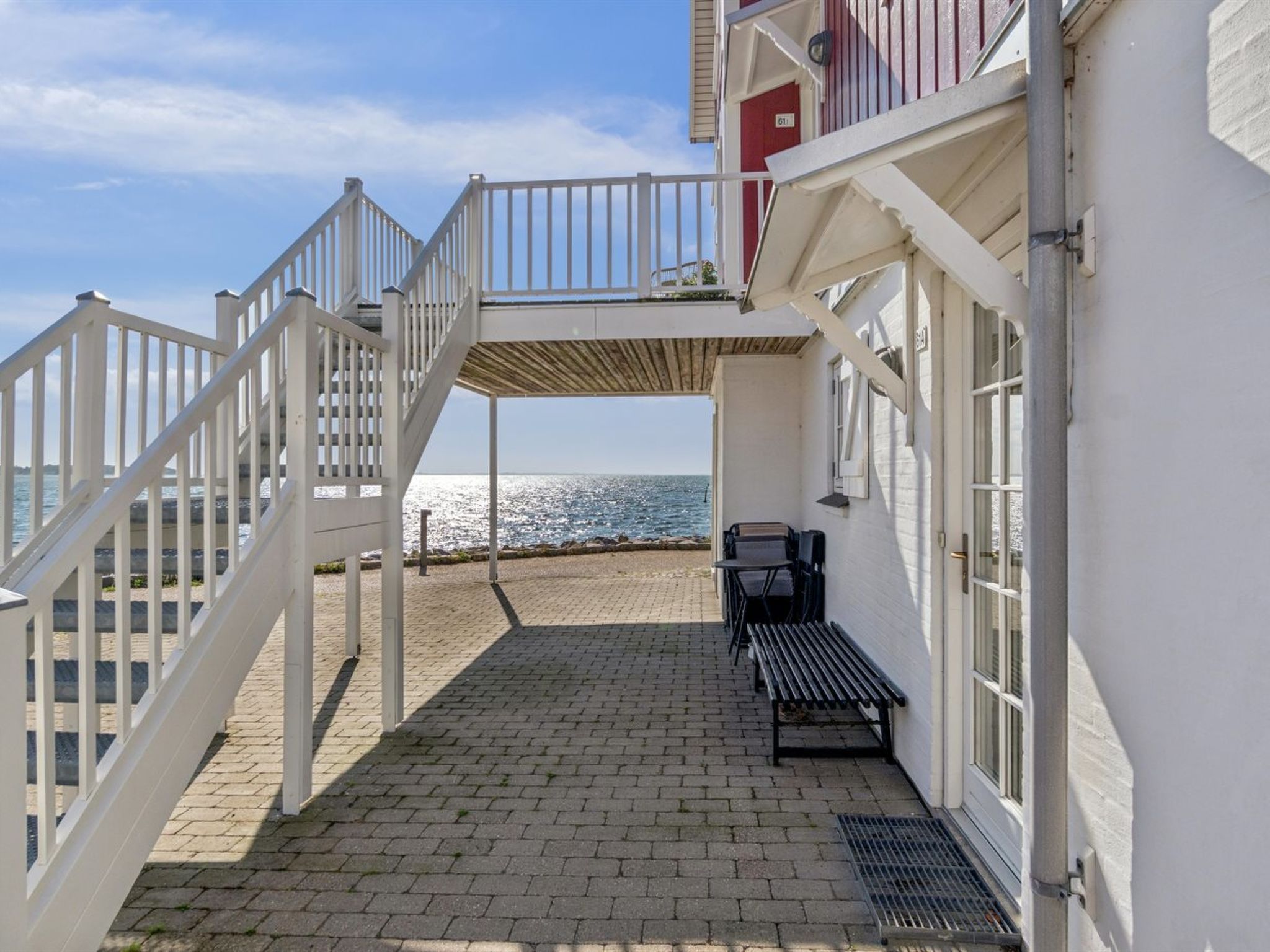 Una propiedad frente al mar con un patio empedrado, un banco de madera y una baranda blanca que da a un balcón con vistas al mar.