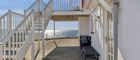 A seaside property with a paved patio, a wooden bench, and a white railing leading to a balcony overlooking the sea.