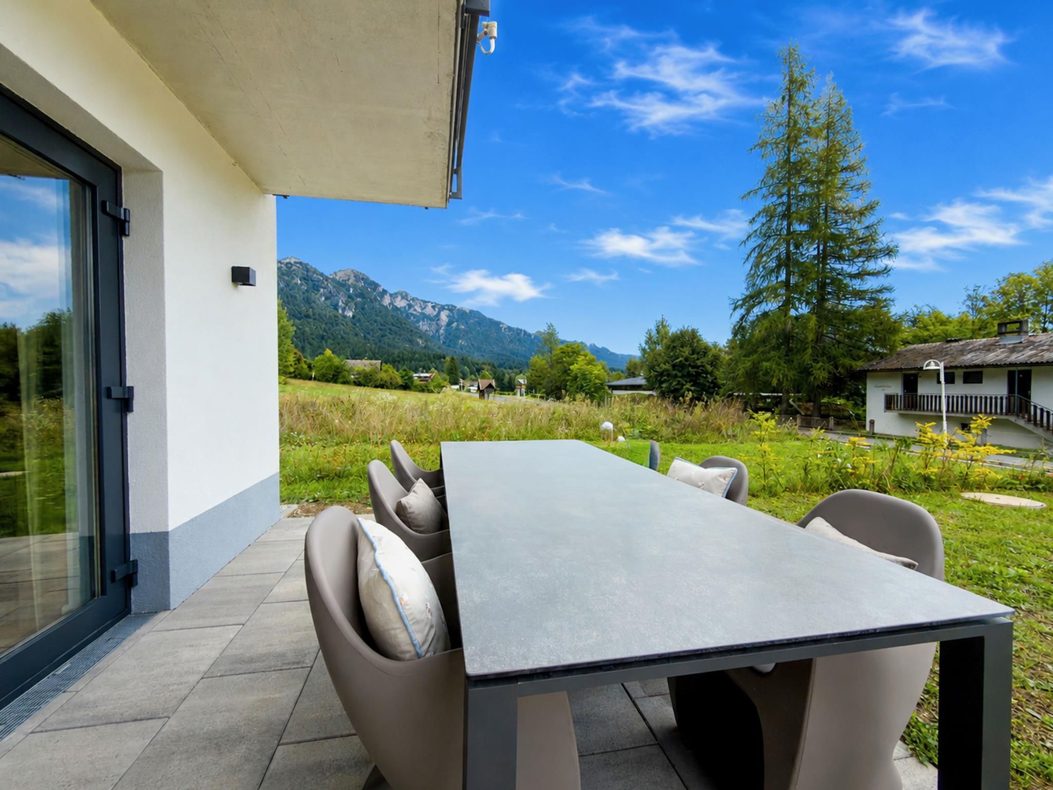 An outdoor dining area with a table and chairs, a glass door, and a view of mountains and trees.