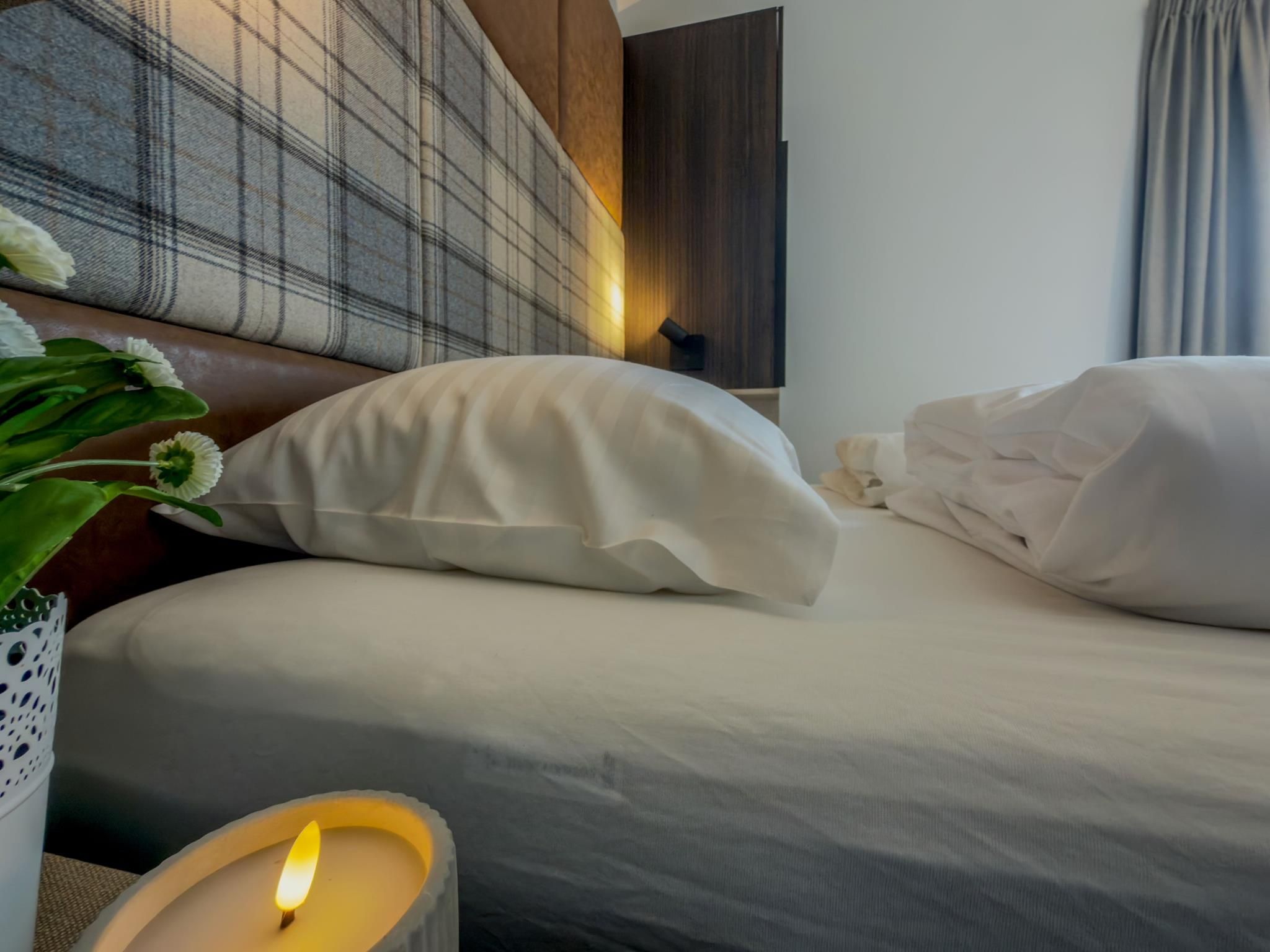 A neatly made bed with white linens and pillows, a lit candle on a bedside table, and a plaid headboard.