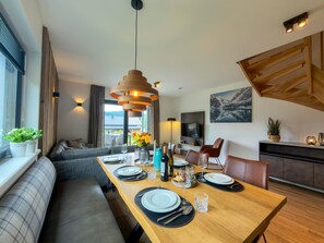 Un espace de repas moderne avec une table en bois dressée pour quatre personnes, un canapé et un téléviseur en arrière-plan.