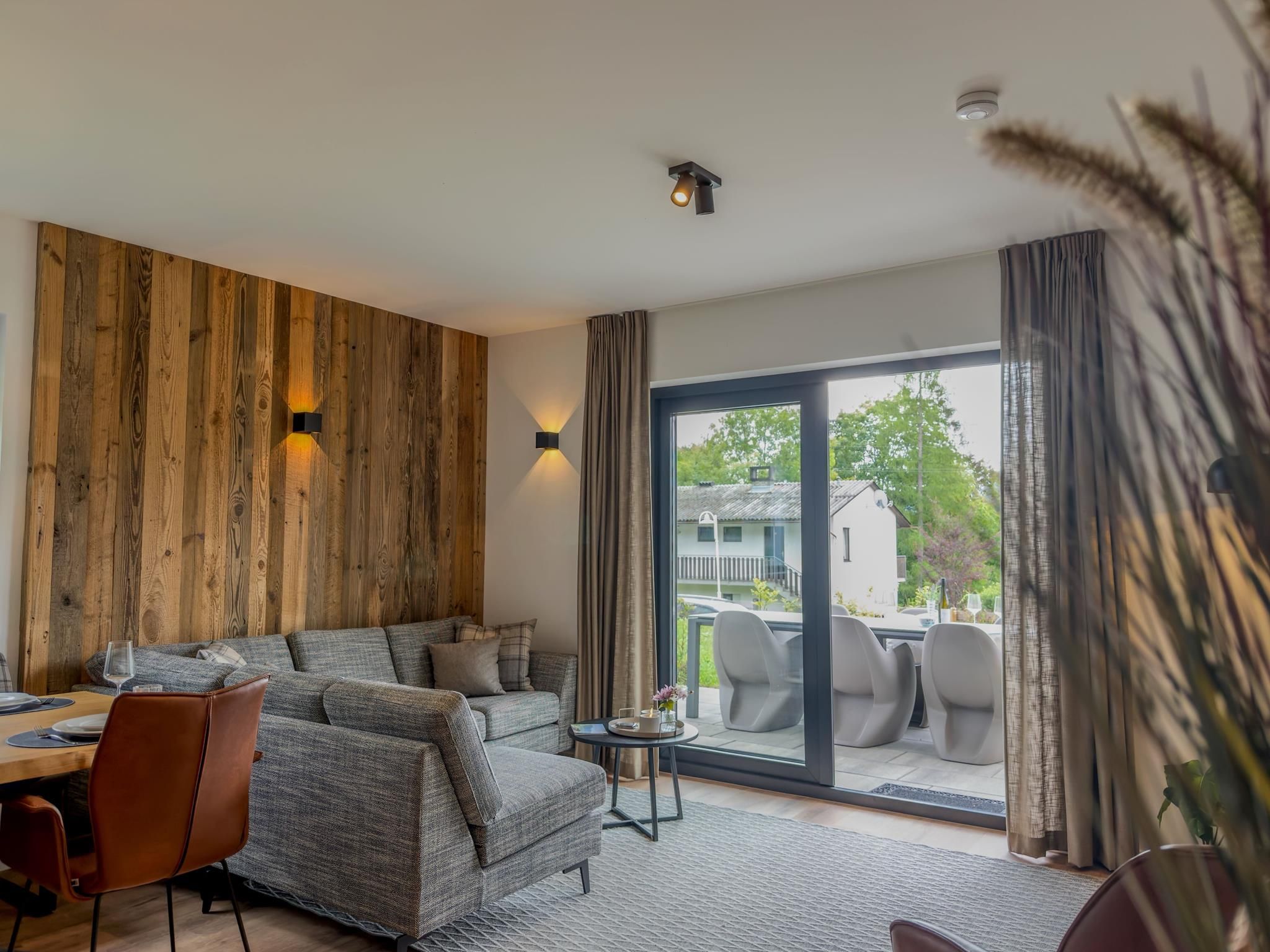 Un salon moderne comprenant un canapé, une table à manger et des chaises, un mur d’accent en bois et une vue dégagée sur l’extérieur grâce à une grande porte-fenêtre coulissante.