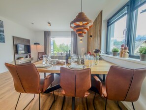 Une salle à manger moderne avec une table en bois dressée pour quatre personnes, des chaises en cuir, un luminaire suspendu et une vue sur l’extérieur grâce à de grandes fenêtres.