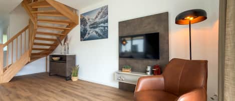 A modern living room with a wooden staircase, a flat-screen TV, a leather chair, and a floor lamp.