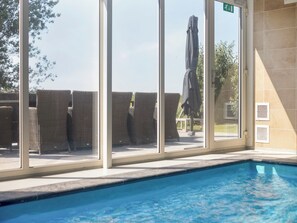 En inomhuspool med väggar av genomskinligt glas, flätade fåtöljer och ett parasoll som kan fällas upp.
