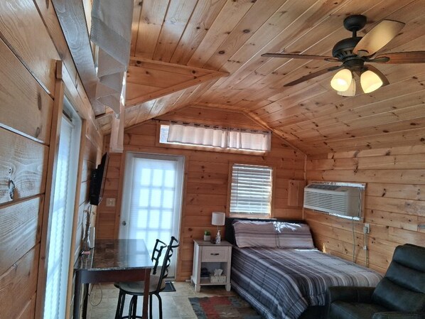 Un dormitorio de cabaña de madera con cama, escritorio, silla, ventilador de techo y ventana con persianas.
