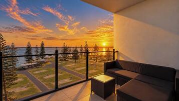A balcony with a view of a sunset over a body of water, surrounded by trees and a residential area.