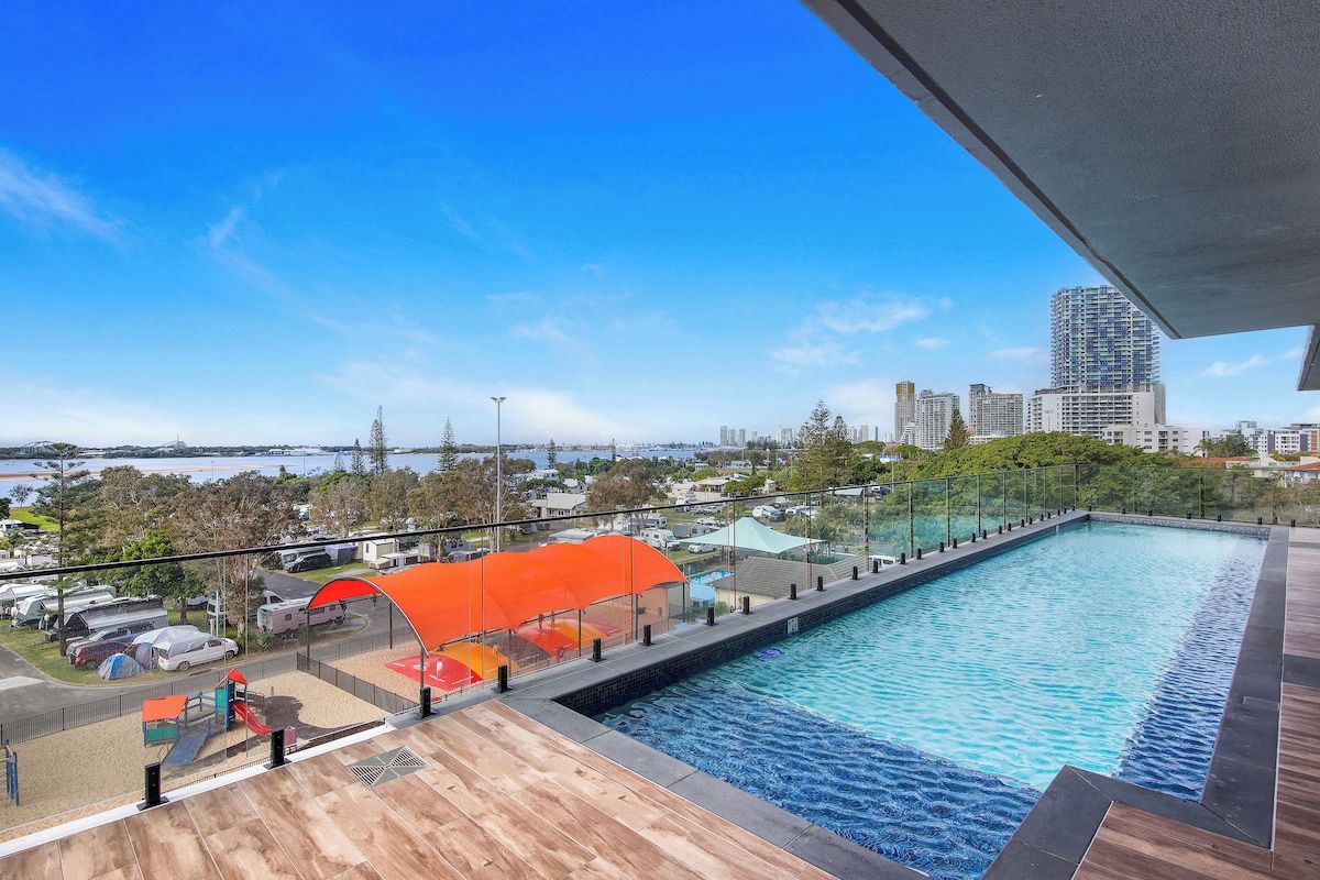 A rooftop swimming pool with a city view, surrounded by a wooden deck and a glass railing.