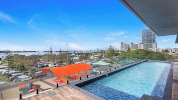 A rooftop swimming pool with a city view, surrounded by a wooden deck and a glass railing.