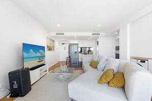 Un salon moderne avec un canapé, une table basse et un téléviseur. On aperçoit à l’arrière-plan un coin cuisine avec un réfrigérateur et un évier.