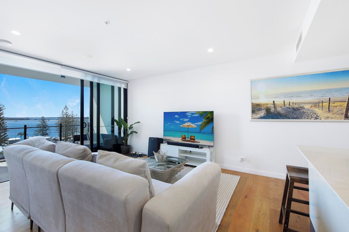 Een moderne woonkamer met een groot raam dat uitkijkt op een wateroppervlak, een flatscreen televisie en een eethoek.
