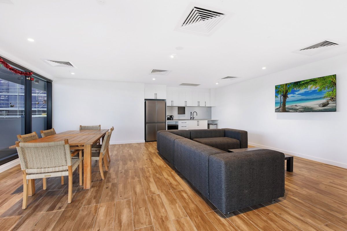 A modern living room with a wooden dining table and chairs, a grey sofa, and a kitchen in the background.