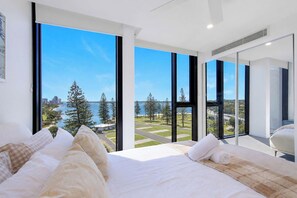 Une chambre moderne avec un grand lit, une literie blanche et une vue sur un paysage côtier grâce à de grandes fenêtres du sol au plafond.