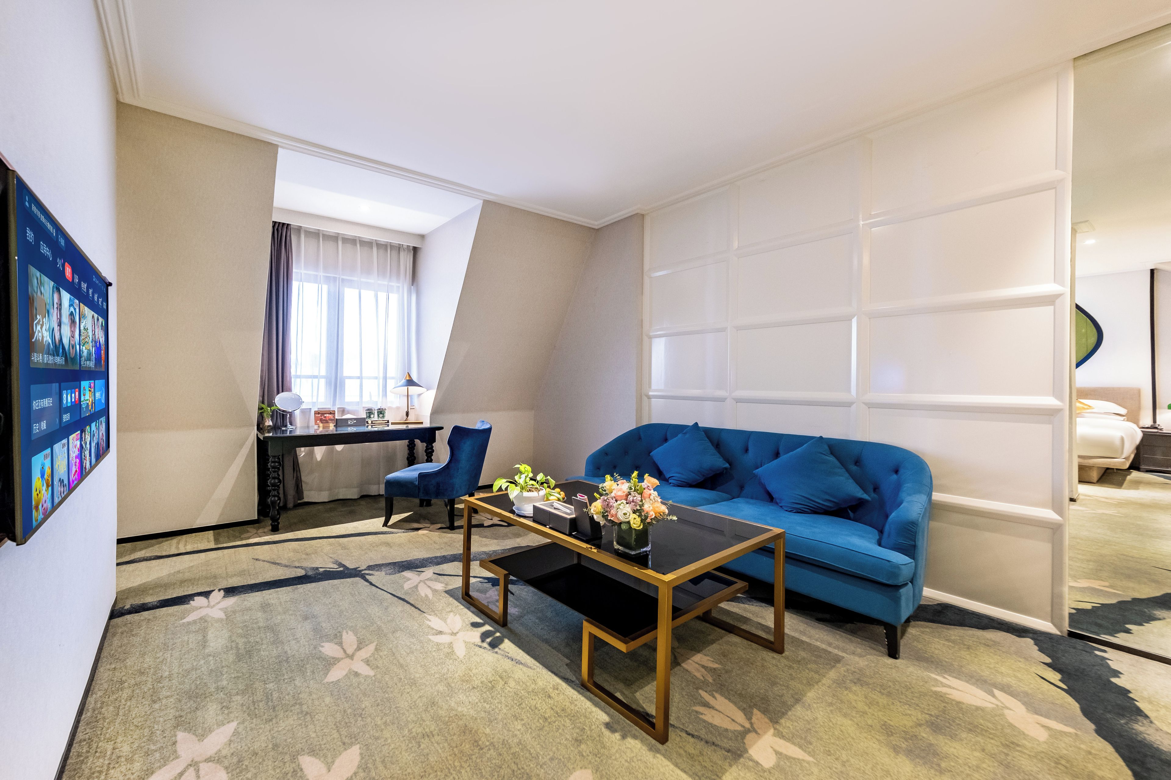 Habitación de hotel con un sofá azul, una mesa de centro, un escritorio con ordenador y un ventanal grande.