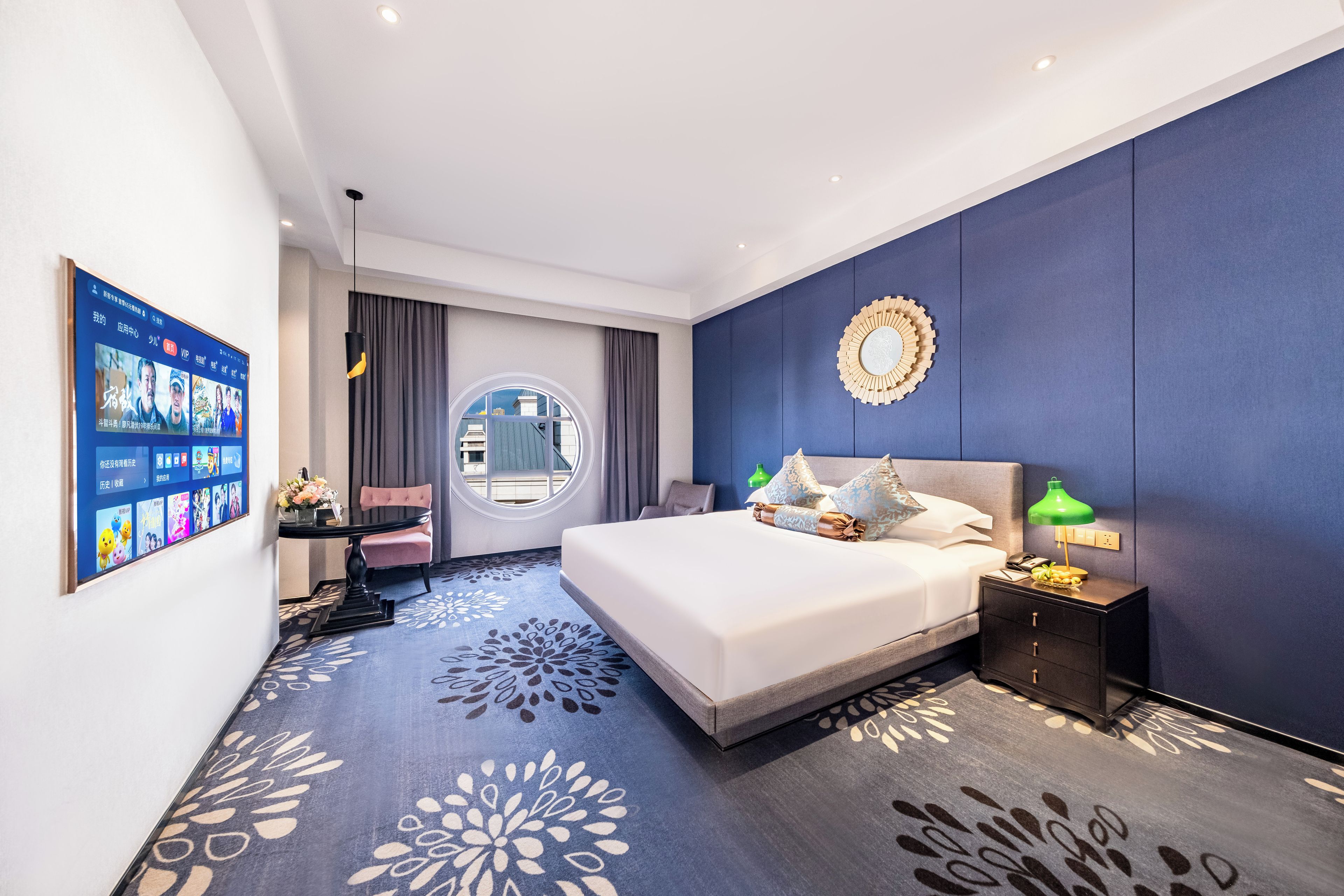 A modern hotel room with a large bed, a round window, a wall-mounted mirror, and a digital screen displaying room information.
