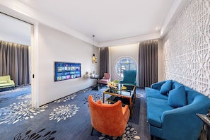 Uma sala de estar moderna com um sofá azul, poltrona laranja e uma mesa redonda. Uma grande tela de TV está fixada na parede, e há uma decoração de parede estampada.