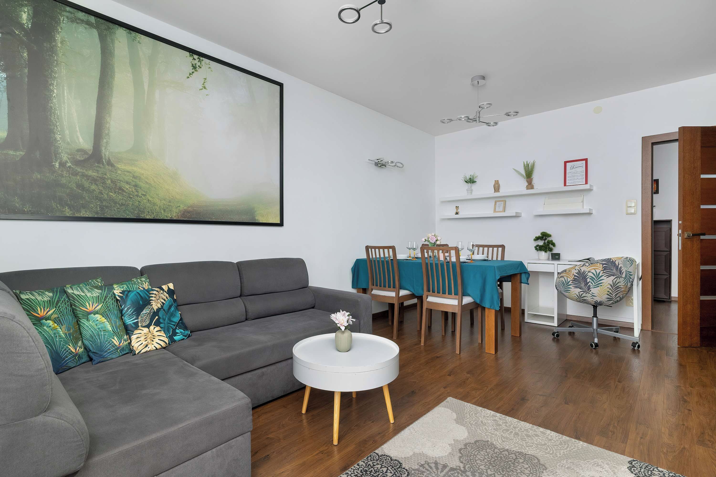 Uma sala de estar moderna com um sofá cinza, uma mesa de centro branca redonda e uma grande pintura com tema de floresta na parede.