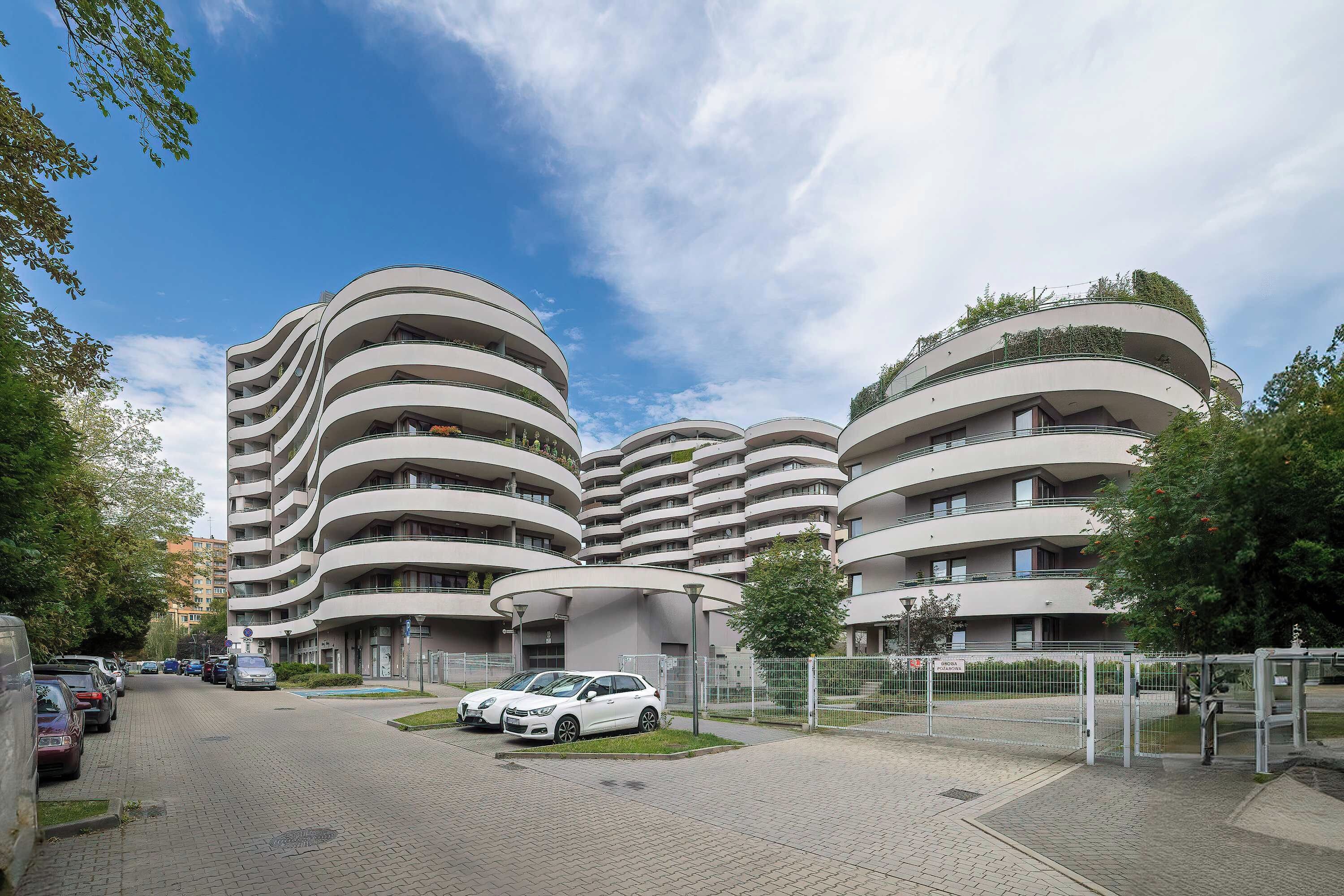 Un bâtiment moderne à plusieurs étages, doté d’une façade incurvée, d’un espace de stationnement avec des voitures et d’un ciel dégagé.