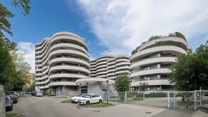 Ein modernes, mehrstöckiges Gebäude mit einer gewölbten Fassade, einem Parkplatz mit Autos und einem wolkenlosen Himmel.