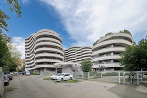 Um edifício moderno de vários andares com fachada curva, área de estacionamento com carros e céu claro.