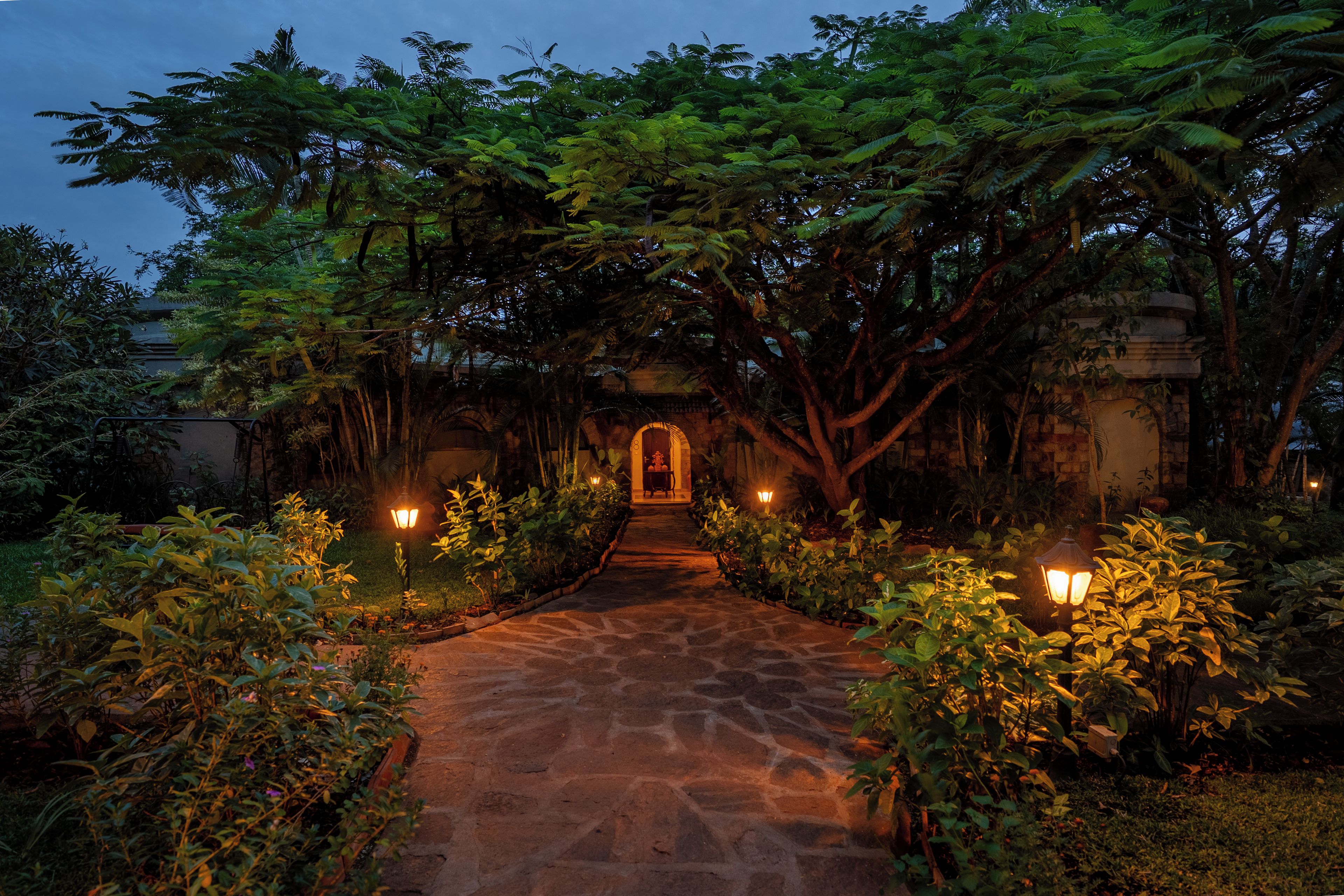 A stone pathway leading to a traditional archway, surrounded by lush greenery and illuminated by wall-mounted lights.