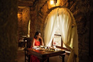 Uma pessoa sentada à mesa de madeira com uma refeição, em um ambiente com paredes de pedra e uma janela com cortinas leves.