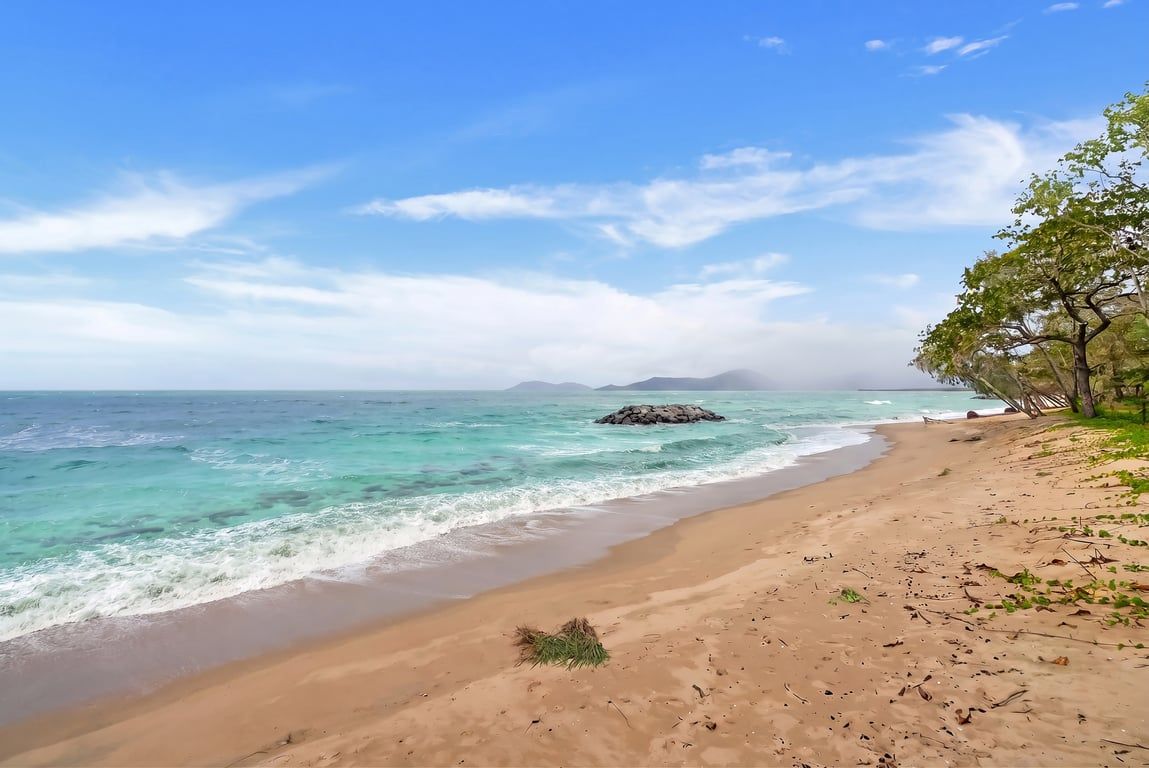 A beach with clear turquoise waters, a rocky outcrop, and a tree-lined path.