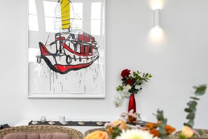 Una obra enmarcada con un barco en la pared, un florero con flores y una mesa con velas.