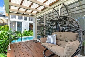 A hanging chair with cushions, a wooden deck, and a glass-enclosed area with a pool in the background. - Belle Escapes Beach Villa San Remo (Holloways Beach)