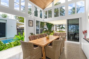Un patio espacioso con una mesa de madera y sillas de mimbre, con vistas a una piscina y abundante vegetación.