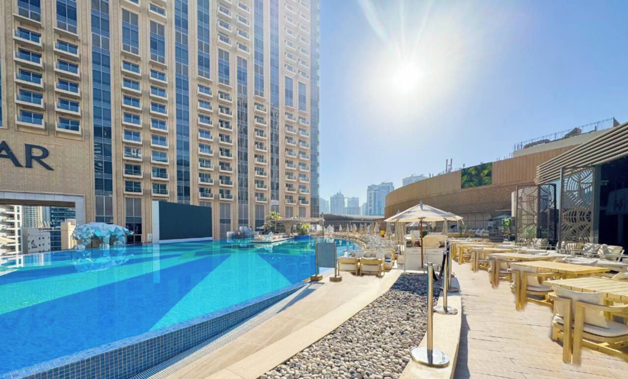 A swimming pool area with a clear sky, a building with a visible 'AR' sign, and a sunlit outdoor seating area.