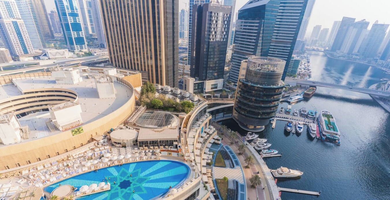 A high-rise hotel with a rooftop pool, a marina with boats, and a cityscape in the background.