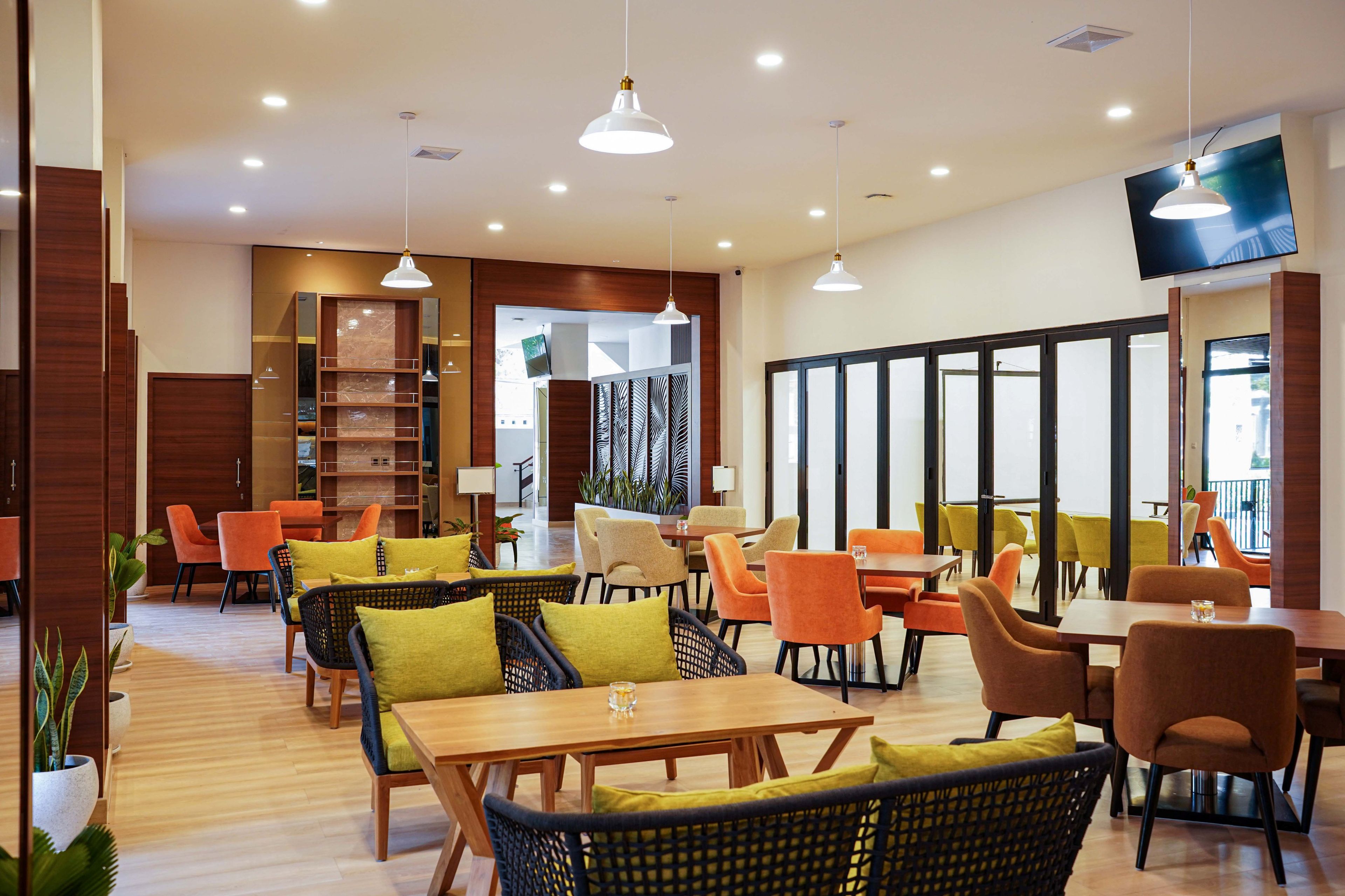 A modern dining area with wooden tables and chairs, pendant lighting, and a large TV screen on the wall.