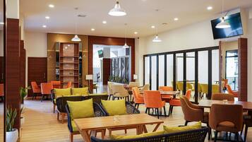 A modern dining area with wooden tables and chairs, pendant lighting, and a large TV screen on the wall.