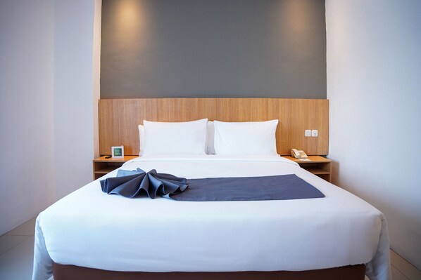 A hotel room with a large bed, wooden headboard, white bedding, a bedside table with a phone, and a wall-mounted light. - Horison Inn Artha Kencana Makassar Hotel (Makassar)