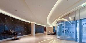 A modern lobby with a curved glass wall, a marble wall with a decorative table, and a seating area with sofas. - The Classy • JW Marriott (Dubai)